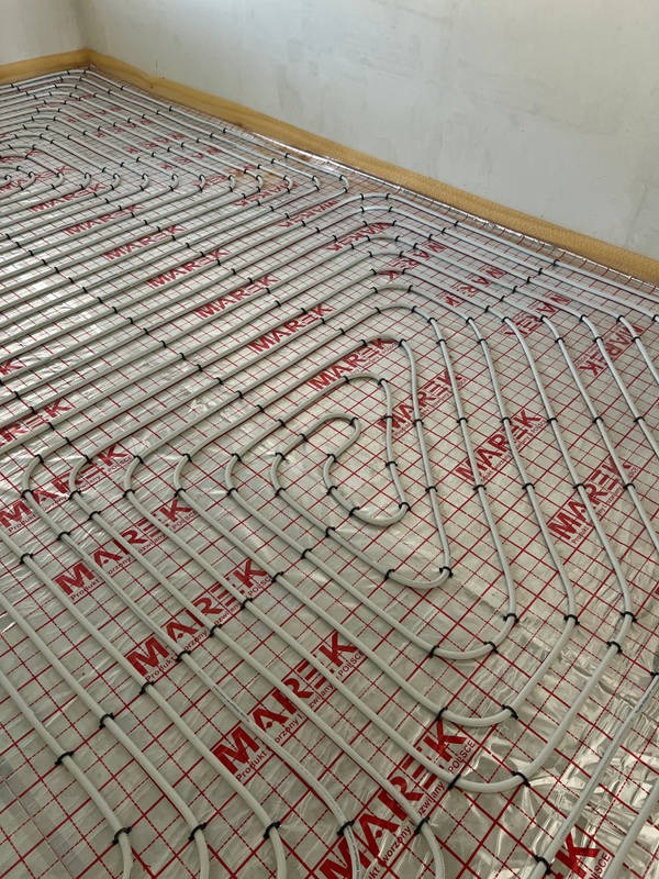 Białe rurki ogrzewania podłogowego ułożone na folii z czerwoną kratką i logotypem producenta, widok z góry, surowy stan ściany w tle.