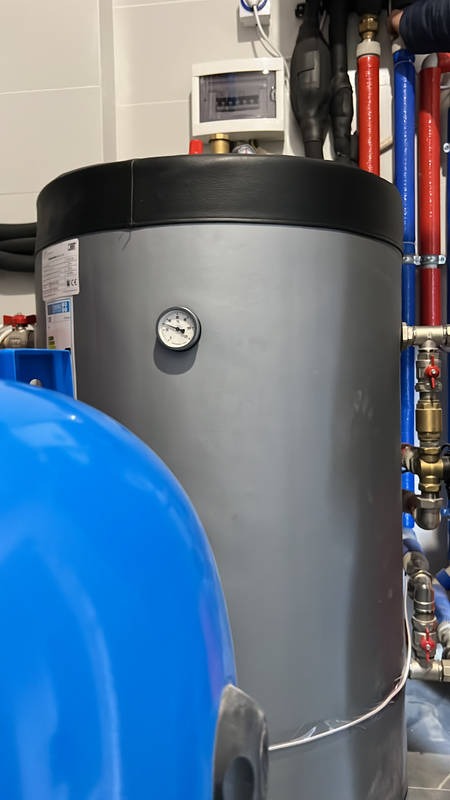 Szary zbiornik buforowy z manometrem, obok niebieskie naczynie przeponowe, widoczne rury instalacji w kolorach czerwonym i niebieskim oraz skrzynka elektryczna na ścianie.
