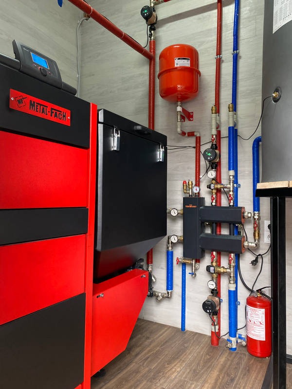 Zainstalowany, nowoczesny piec Metal-Fach z podajnikiem, widoczne rury instalacji grzewczej w kolorach czerwonym i niebieskim oraz zbiornik wyrównawczy i gaśnica.
