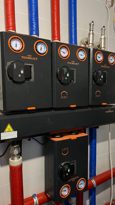 Cztery czarne moduły pompy ciepła Termojet z pomarańczowymi akcentami, manometrami i wyświetlaczami, zamontowane na ścianie z widocznymi rurami w izolacji czerwonej i niebieskiej.