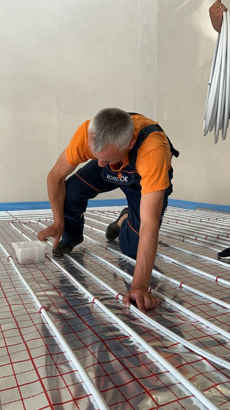 Instalator w kombinezonie roboczym układa białe rurki ogrzewania podłogowego na folii z czerwoną siatką, przymocowując je plastikowymi elementami.