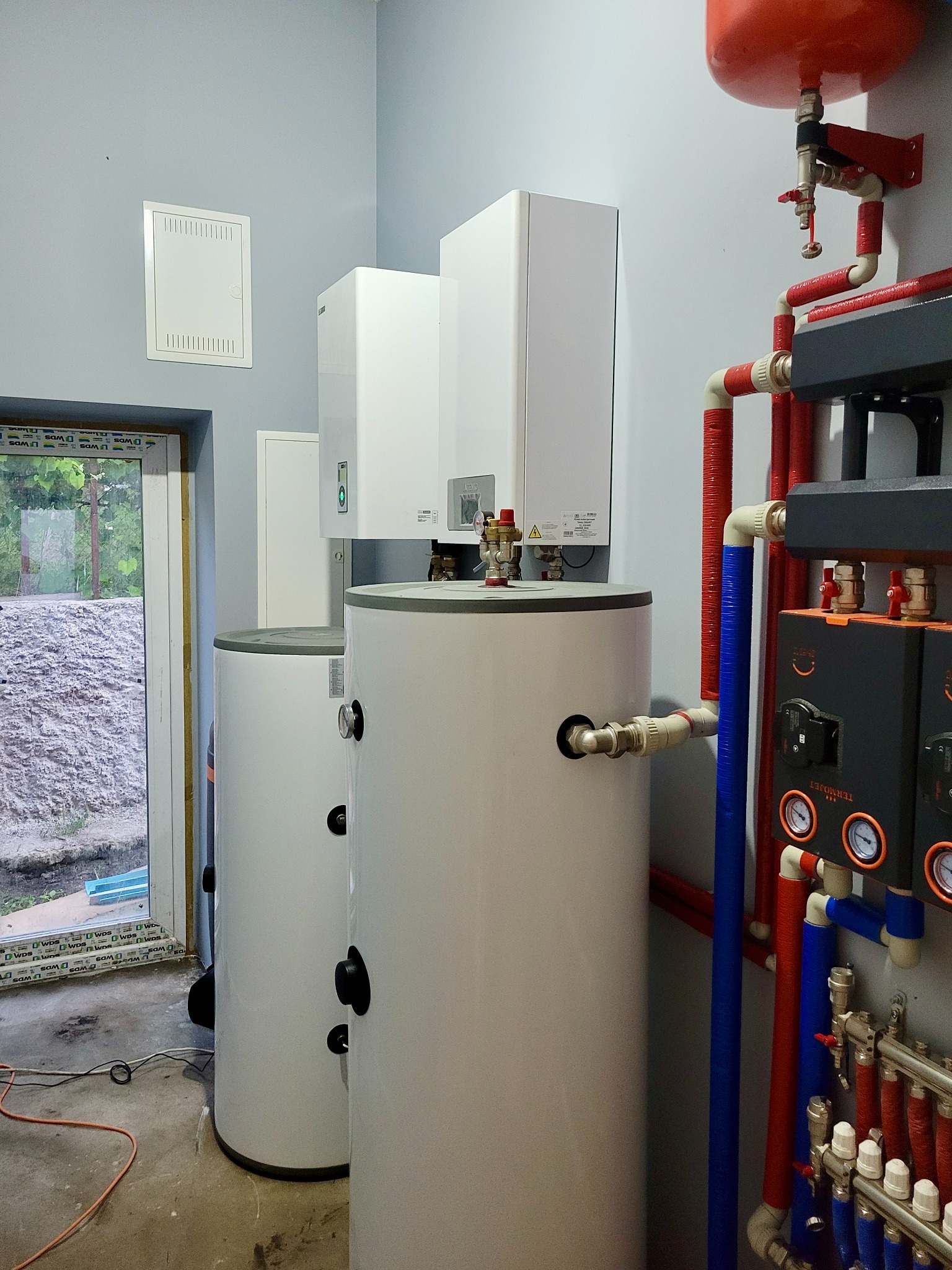 Dwa białe zbiorniki akumulacyjne ciepła w kotłowni, z widocznymi rurami w kolorach czerwonym i niebieskim, oraz panelem sterowania z manometrami, na tle szarej ściany i okna z widokiem na ziemię.