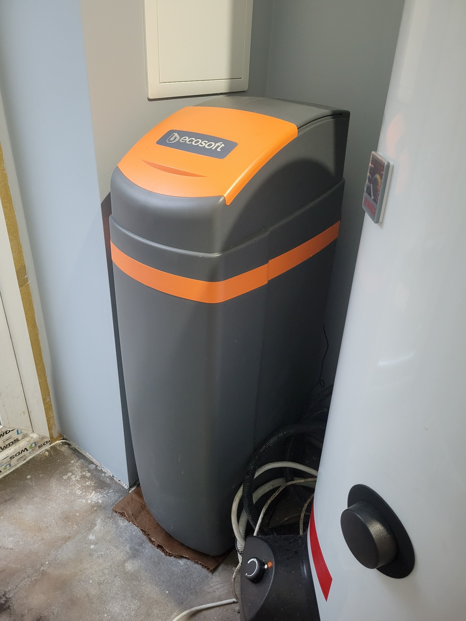 Szaro-pomarańczowy zmiękczacz wody Ecosoft stojący obok białego zasobnika ciepłej wody w pomieszczeniu gospodarczym z szarą ścianą i brudną podłogą.