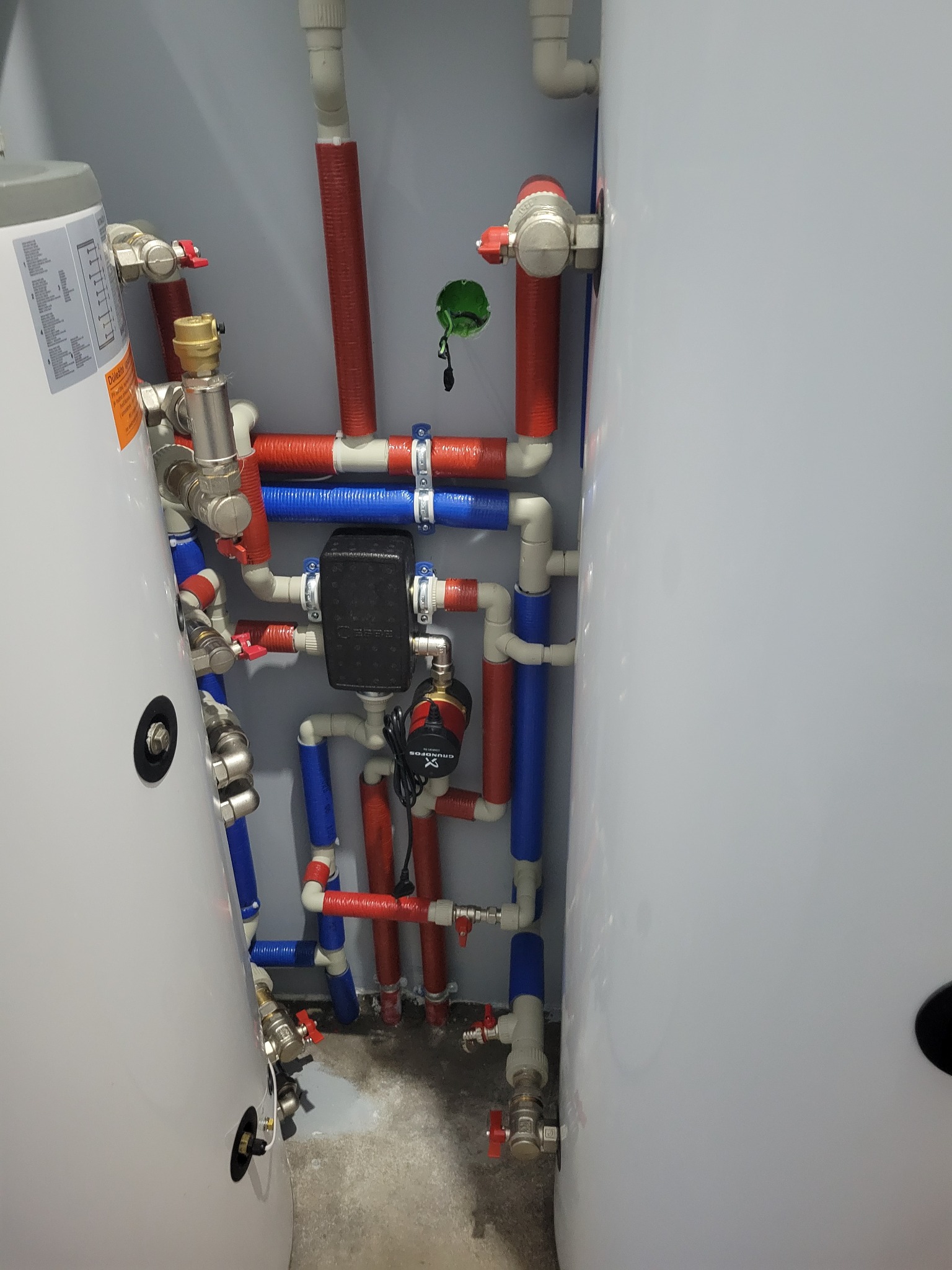 Kompleksowa instalacja grzewcza z dwoma zasobnikami ciepłej wody, rurami w izolacji czerwonej i niebieskiej, pompą obiegową Grundfos i automatyką, widok z bliska.