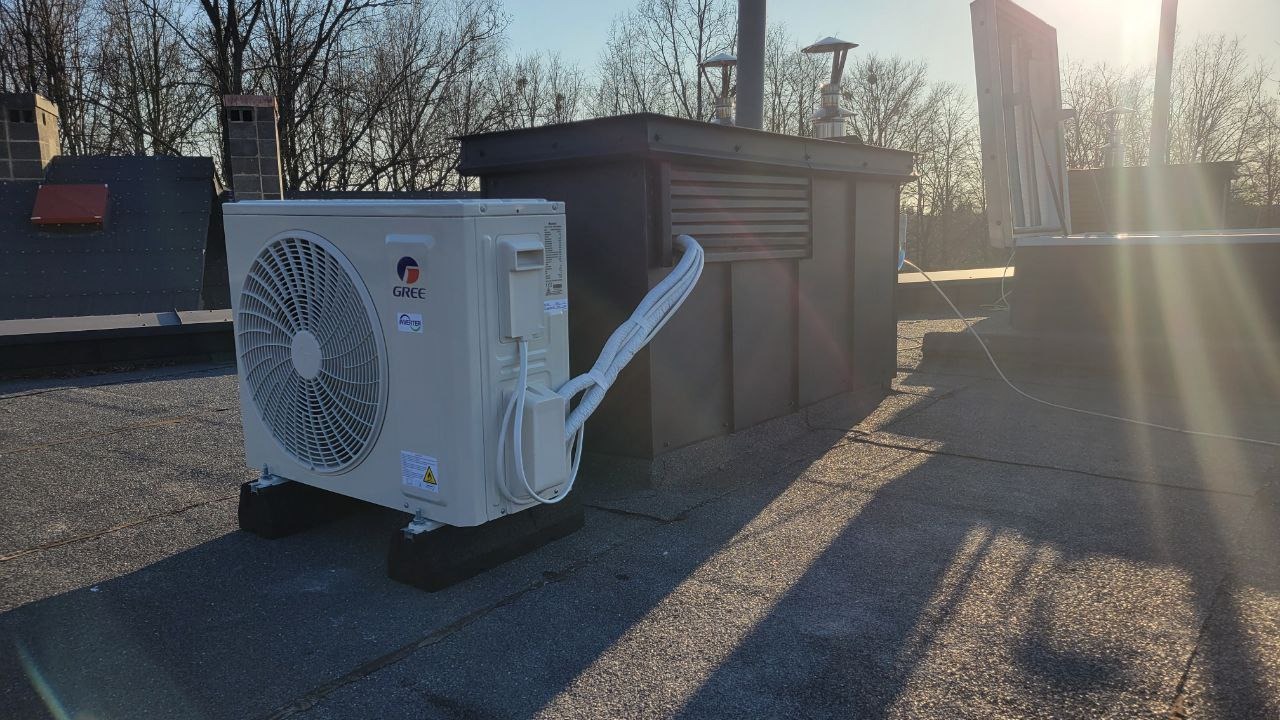 Biała jednostka zewnętrzna klimatyzatora marki Gree, umieszczona na czarnych gumowych podkładkach na płaskim dachu, z widocznymi przewodami łączącymi ją z metalową obudową wentylacyjną w tle,...