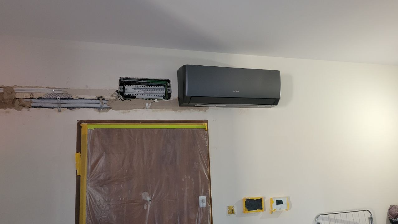 Szara jednostka klimatyzacji zamontowana na ścianie w trakcie remontu, widoczne kable i rury w ścianie, zabezpieczone drzwi folią malarską.
