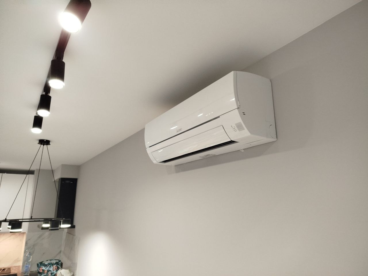 Biały klimatyzator Mitsubishi Electric zamontowany na szarej ścianie w nowoczesnym wnętrzu, widoczna lampa sufitowa z reflektorami.