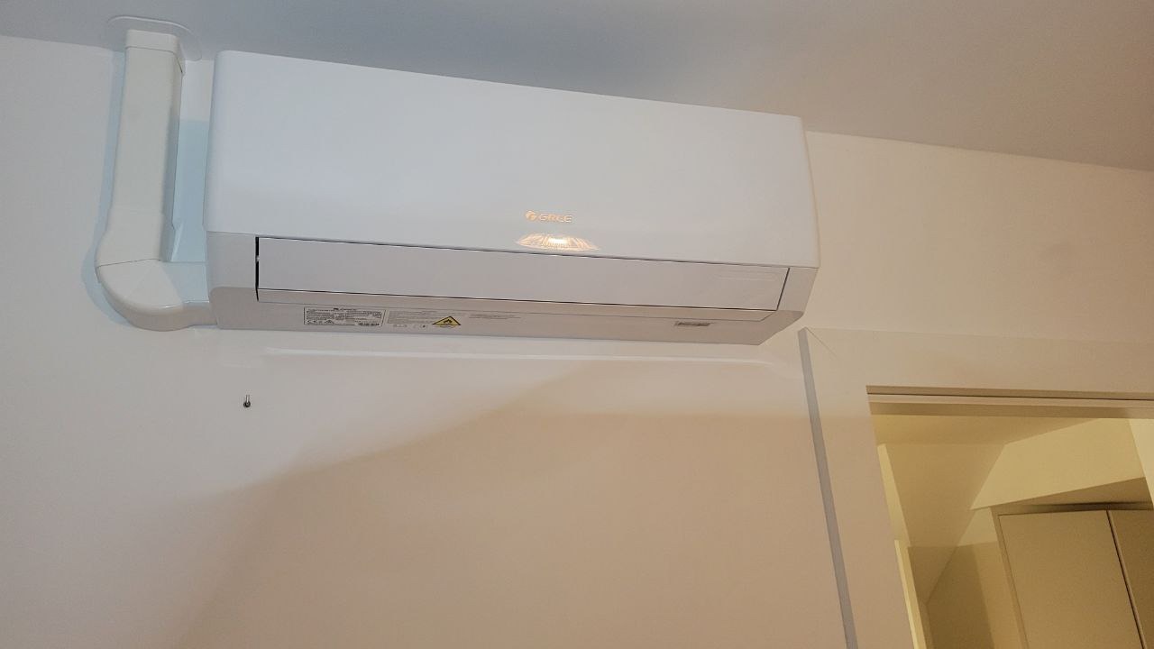 Biały klimatyzator wiszący marki Gree zamontowany na ścianie w jasnym pomieszczeniu, widoczny fragment instalacji odprowadzającej skropliny i narożnik futryny drzwiowej.