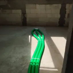 Trzy zielone rury wentylacyjne, spięte białą taśmą, leżą na szarej betonowej podłodze w surowym wnętrzu, z widocznym oknem dachowym i fragmentami ściany z bloków.