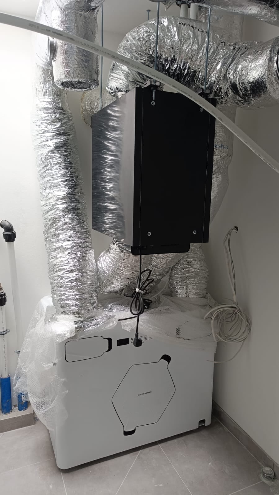 Nowoczesna centrala wentylacyjna z rekuperacją, jeszcze w folii ochronnej, podłączona do srebrnych, elastycznych przewodów wentylacyjnych, zamontowana w jasnym pomieszczeniu.