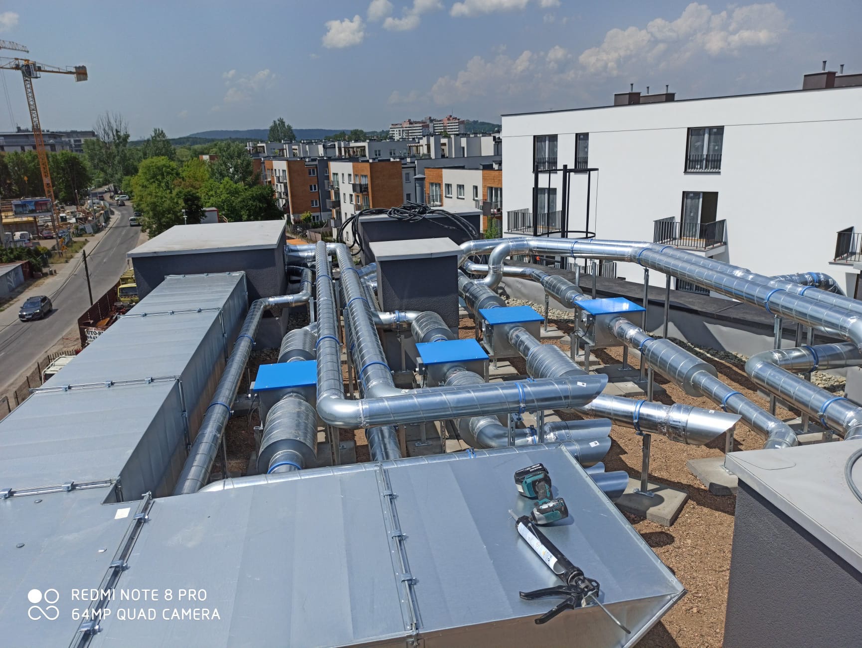 System kanałów wentylacyjnych z niebieskimi elementami na dachu budynku, z narzędziem do uszczelniania i wiertarką akumulatorową na pierwszym planie, w tle widok na miasto i budowę.