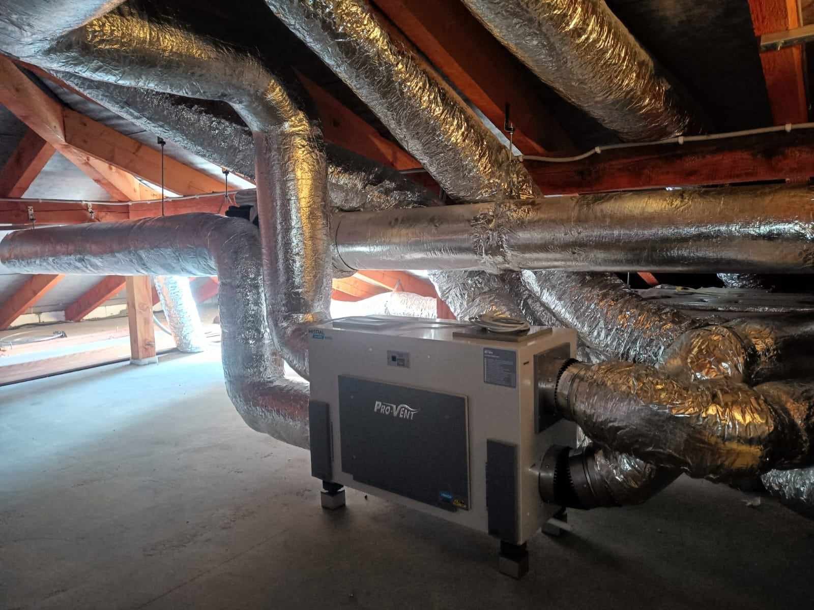 Centrala wentylacyjna Pro-Vent z podłączonymi przewodami wentylacyjnymi w folii aluminiowej, zamontowana na poddaszu o drewnianej konstrukcji dachowej.