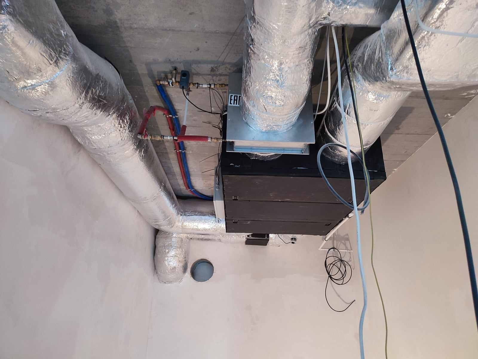 Widok z dołu na zamontowaną instalację rekuperacji z aluminiowymi rurami, zaworami i przewodami pod sufitem w surowym stanie.