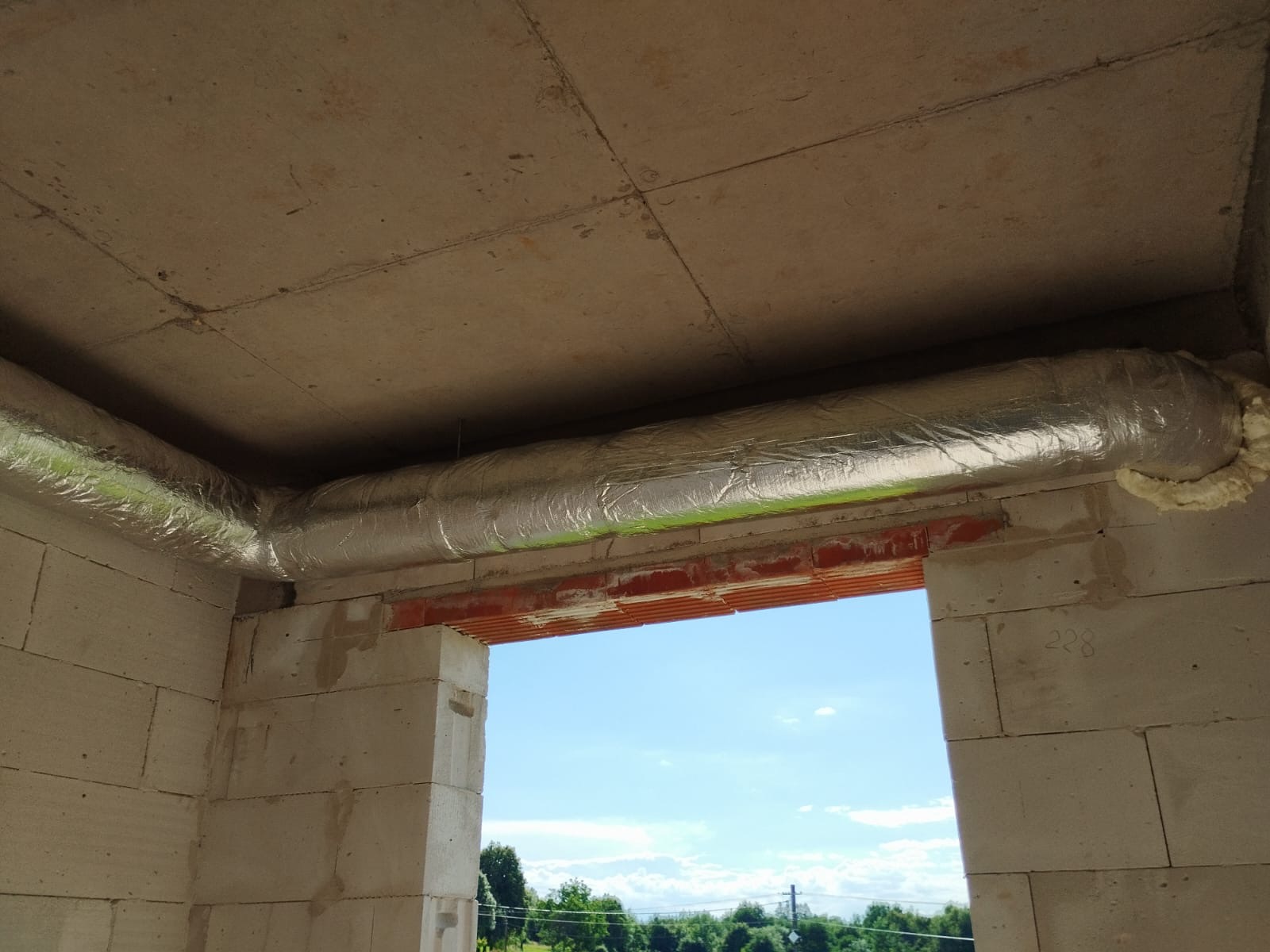 Fragment surowego wnętrza z izolowanym przewodem wentylacyjnym pod sufitem, widok przez otwór okienny na zewnątrz.