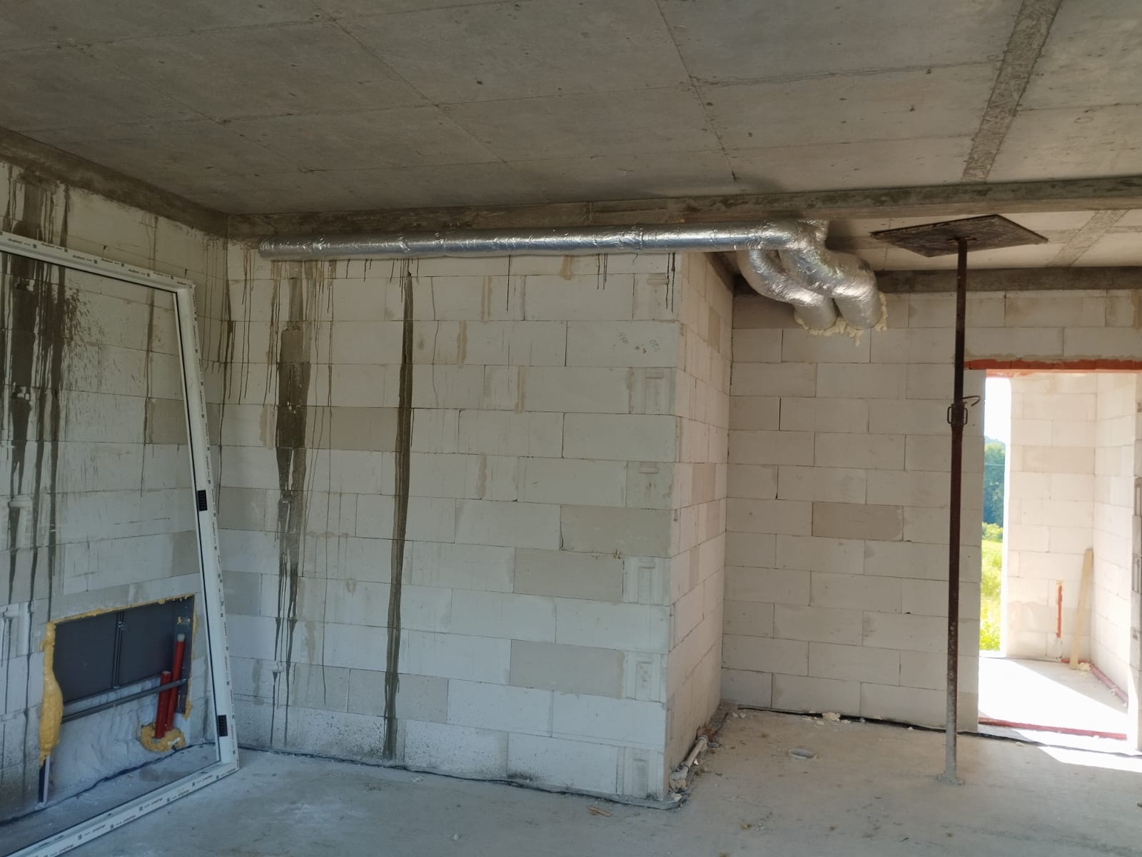 Wnętrze surowego budynku z widoczną instalacją wentylacyjną z izolowanych rur aluminiowych pod sufitem, ściany z białych bloczków, widoczne ślady po zalaniu.