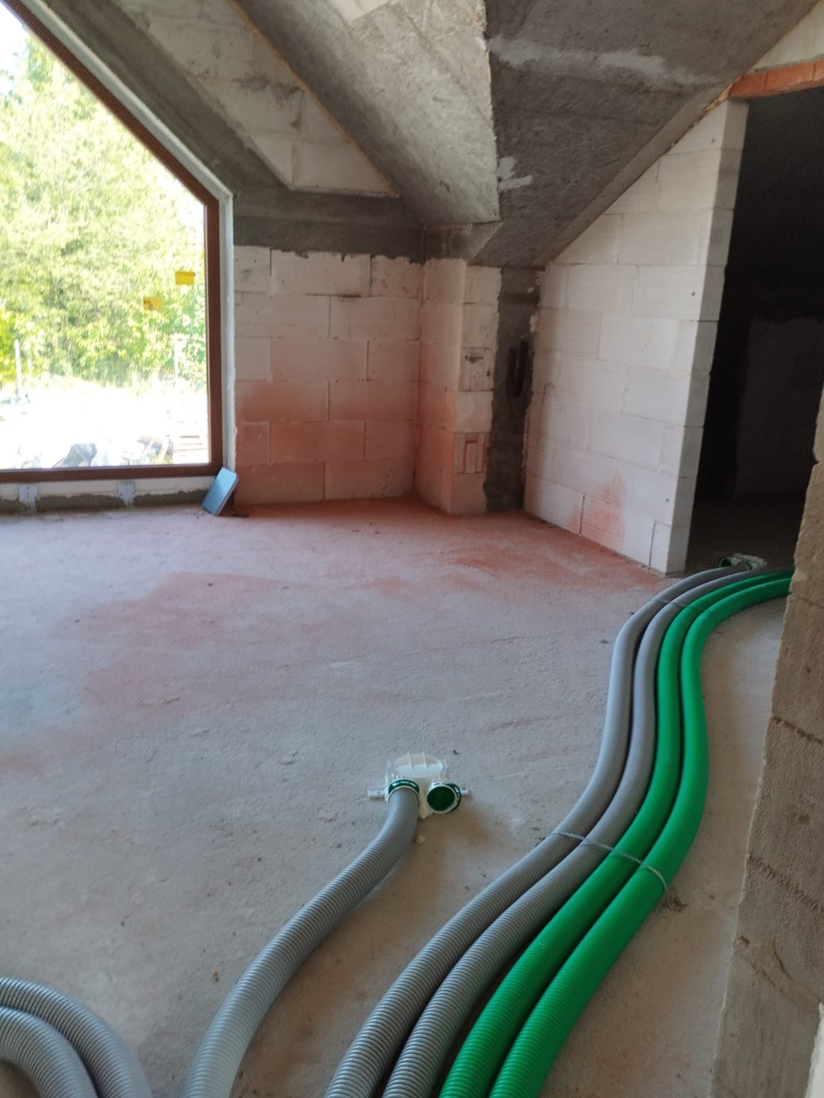 Surowe wnętrze w trakcie budowy z widocznymi szarymi i zielonymi rurami wentylacyjnymi na podłodze, okno z widokiem na zieleń oraz surowe ściany i strop.