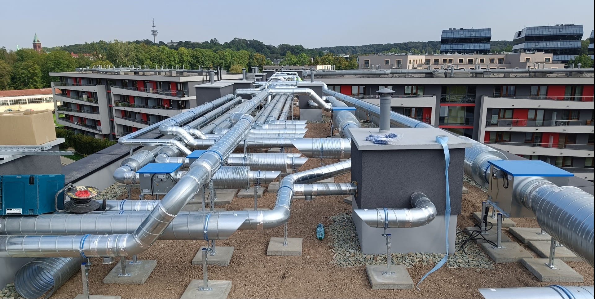 Rozbudowany system srebrnych rur wentylacyjnych na dachu budynku mieszkalnego, widoczne elementy mocujące, niebieskie skrzynki i budynki w tle.