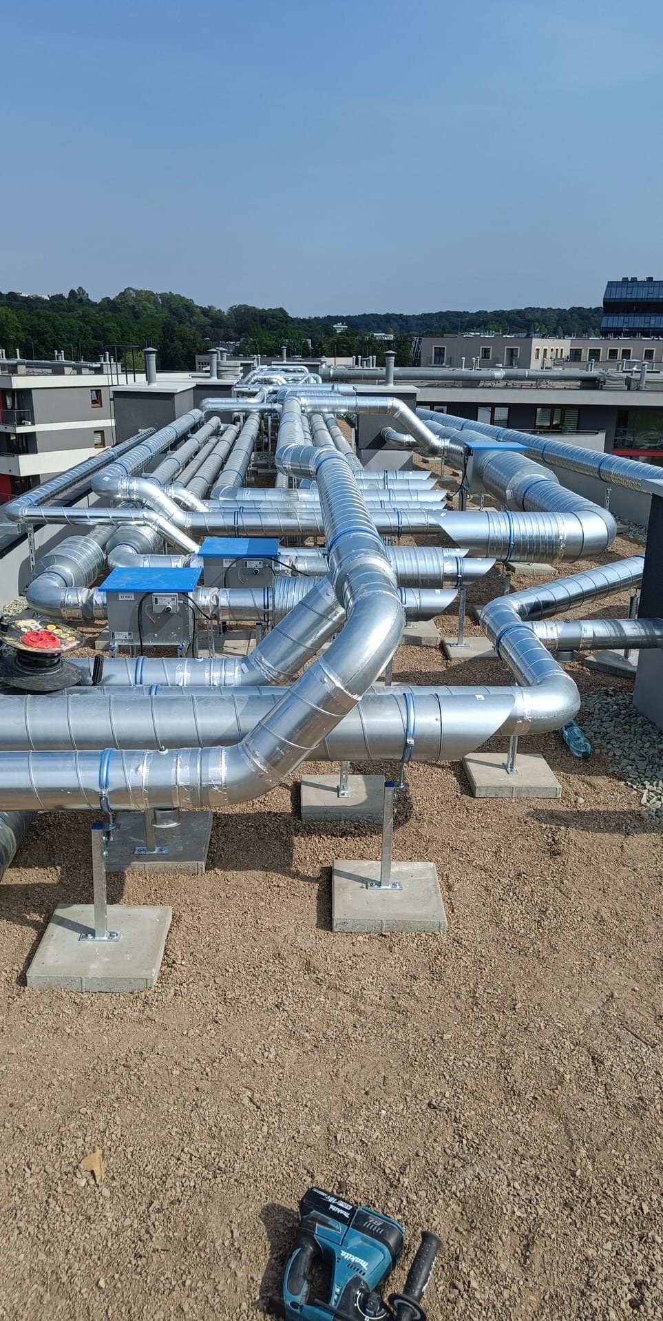 Widok z góry na rozbudowany system srebrnych rur wentylacyjnych na dachu budynku, wspartych na betonowych podstawach, z narzędziem akumulatorowym w prawym dolnym rogu.