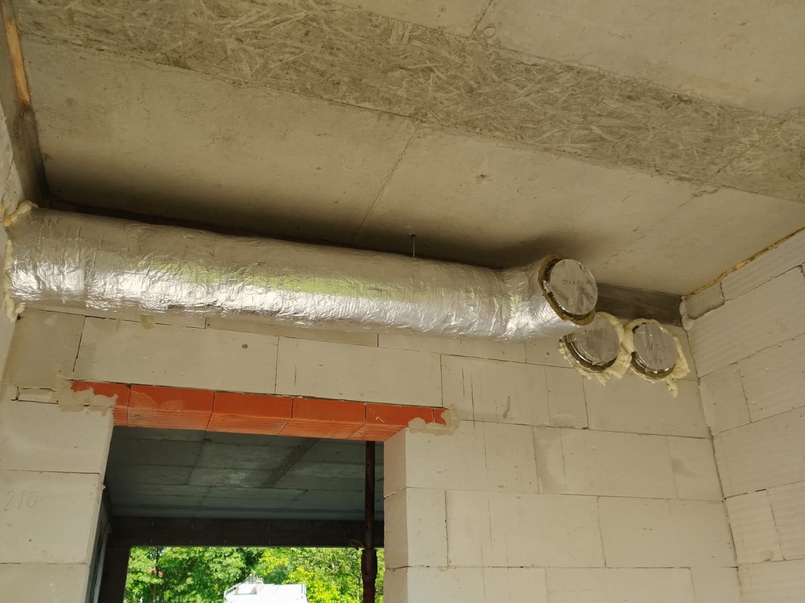 Izolowany, aluminiowy przewód wentylacyjny zamontowany pod stropem w surowym wnętrzu, widoczne trzy okrągłe wyloty zabezpieczone pianką montażową, ściany z białego betonu komórkowego, niewykończony...