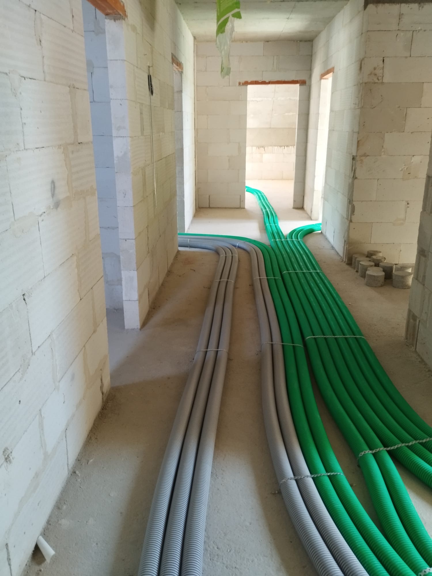 Wnętrze surowego budynku w trakcie instalacji przewodów wentylacyjnych: szare i zielone rury prowadzone wzdłuż ścian z betonu komórkowego, widoczne otwory drzwiowe.