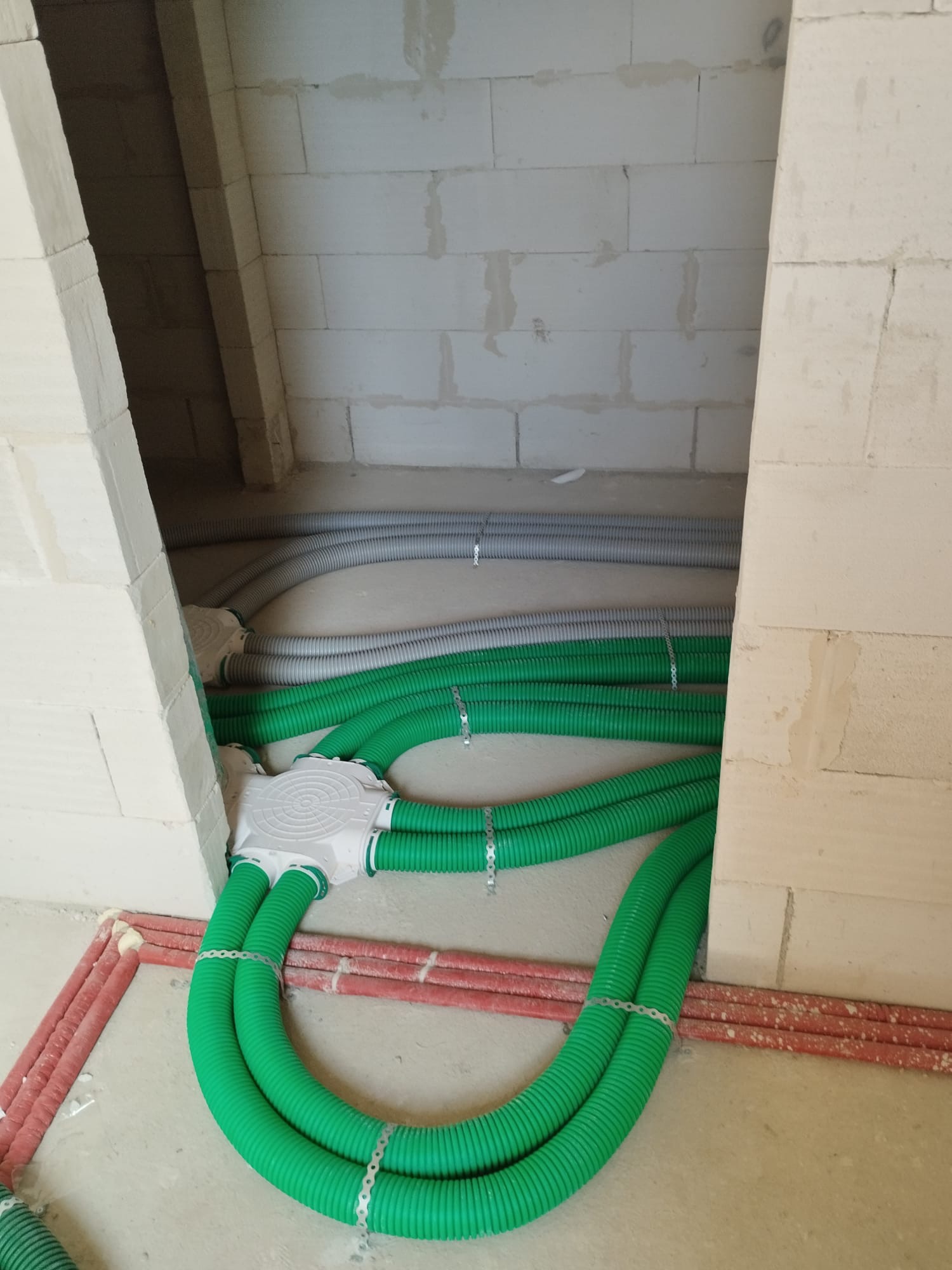 Instalacja przewodów wentylacyjnych w surowym wnętrzu: zielone i szare rury karbowane przymocowane do ściany z betonu komórkowego za pomocą metalowych obejm.