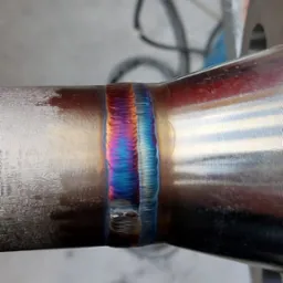 Spoina spawalnicza na metalowej rurze, z widocznymi kolorami tęczy wskazującymi na temperaturę spawania, w tle warsztat z przewodami i rurami PCV.