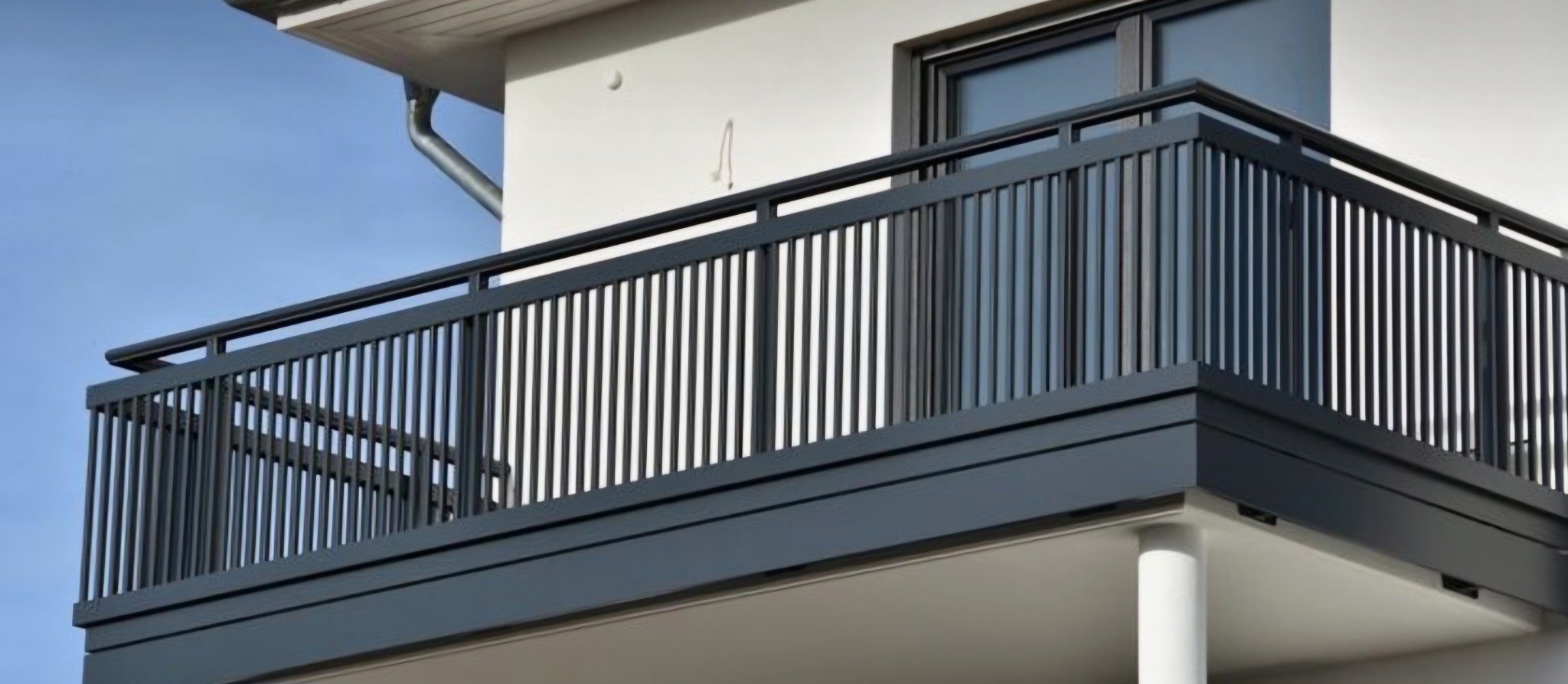 Nowoczesna balustrada balkonowa z pionowymi, metalowymi szczeblami w kolorze antracytowym, zamontowana na balkonie z widokiem na błękitne niebo.