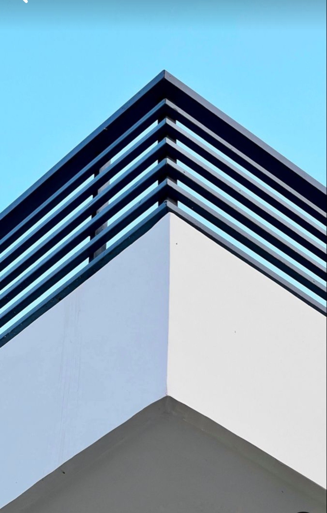 Minimalistyczne ujęcie narożnika budynku z metalową balustradą o poziomym układzie szczebli na tle błękitnego nieba.