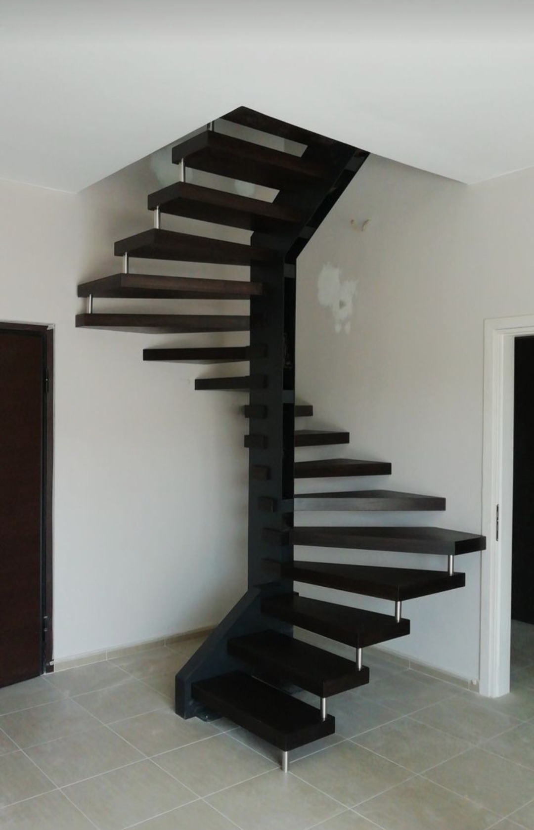 Wewnętrzne schody spiralne z ciemnego drewna na metalowej konstrukcji, wsparte na metalowych nogach, w jasnym pomieszczeniu z szarą podłogą z płytek.