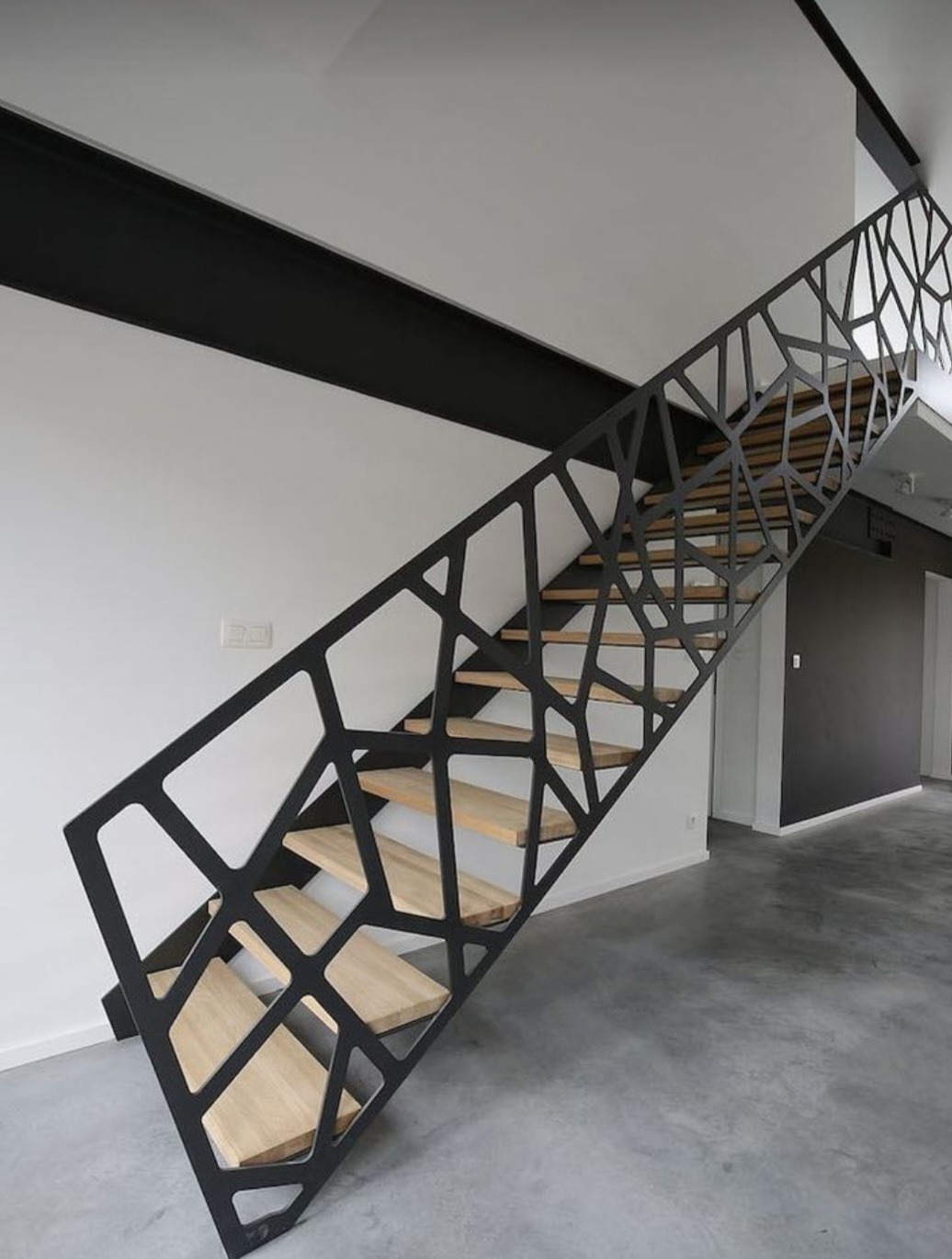 Nowoczesne schody z drewnianymi stopniami i czarną, ażurową balustradą o nieregularnym wzorze, widoczne z dołu pod kątem, na tle białej ściany i szarej betonowej podłogi.