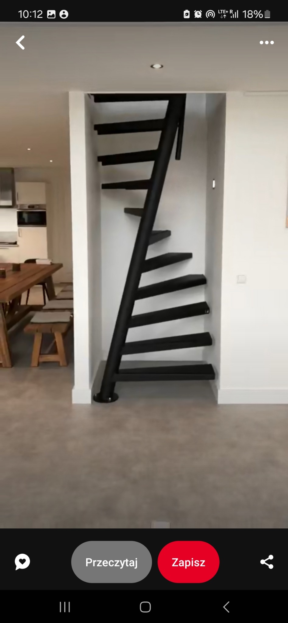 Minimalistyczne, czarne schody spiralne z metalową konstrukcją, wkomponowane w białą wnękę w nowoczesnym wnętrzu, widoczne z perspektywy dolnej, z fragmentem drewnianego stołu i krzeseł w tle.