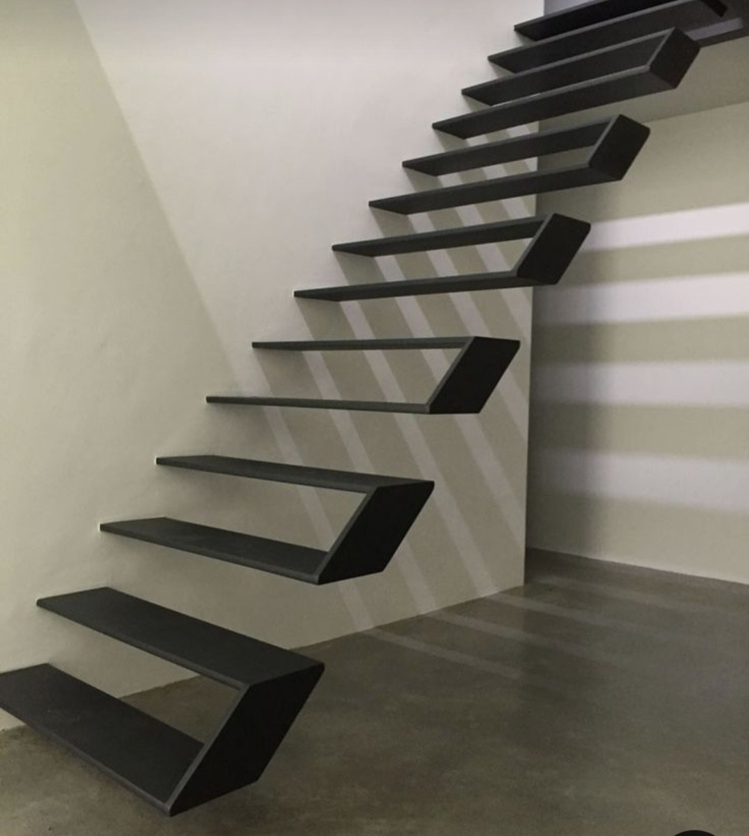 Nowoczesne, minimalistyczne schody wspornikowe z metalu, z geometrycznym kształtem stopni, zamontowane przy jasnej ścianie. Stopnie w kolorze grafitowym, tworzące efekt lewitacji. Widoczny betonowy...