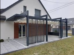 Nowoczesna pergola z metalową konstrukcją i rozsuwanym zadaszeniem materiałowym, przylegająca do jasnego domu z brązową elewacją, widoczna z boku na tle szarej kostki brukowej i trawnika.