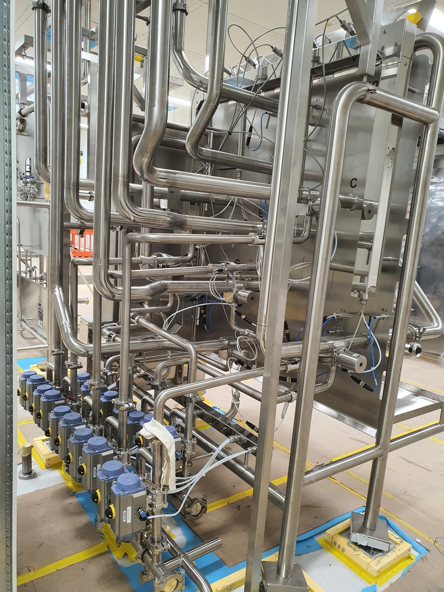 Kompleksowa instalacja z rur ze stali nierdzewnej, z licznymi zaworami i przewodami, zamontowana na ramie, widoczna w hali produkcyjnej.