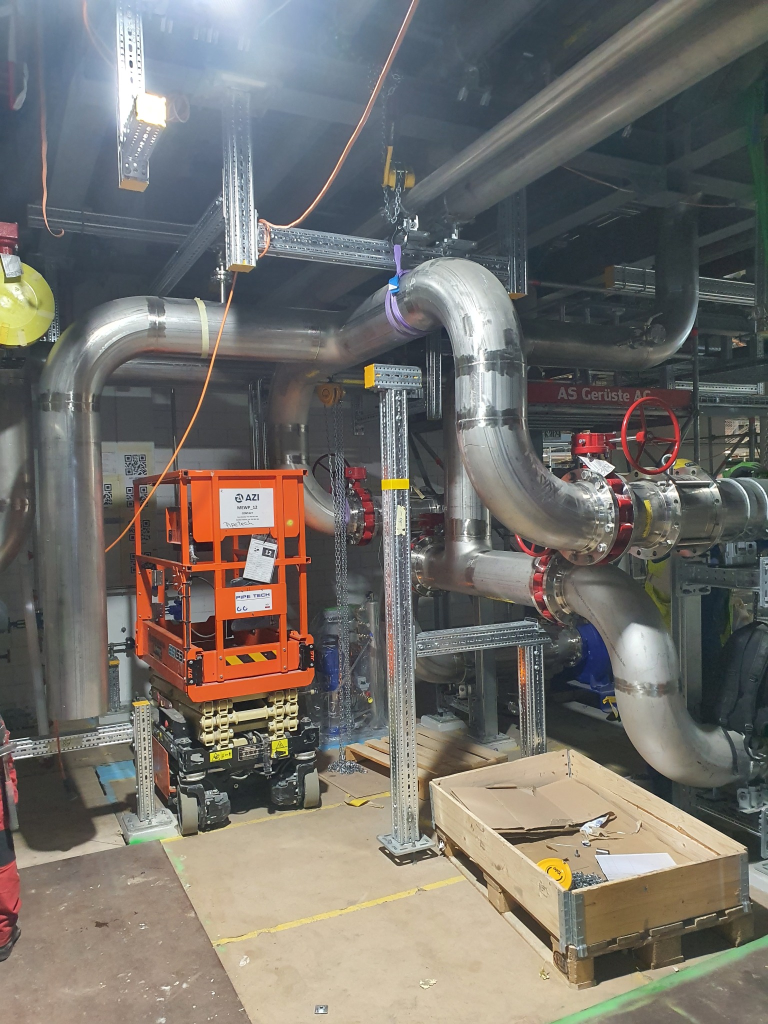 Montaż przemysłowej instalacji rurociągowej z wykorzystaniem podnośnika nożycowego i konstrukcji wsporczych, widoczne rury ze stali nierdzewnej, zawory z czerwonymi pokrętłami i skrzynia...