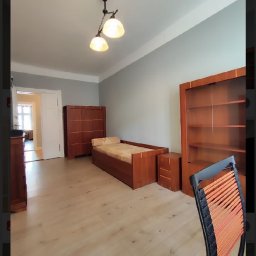 Mariusz Lyczewski - Jasne, świeżo odmalowane wnętrze pokoju z drewnianymi meblami: łóżkiem, szafą i regałem, podłoga z paneli, widok na kolejne pomieszczenie przez otwarte drzwi.