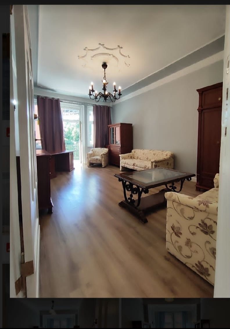 Jasne wnętrze pokoju dziennego z drewnianą podłogą, meblami w stylu retro i żyrandolem, widok zza uchylonych drzwi.