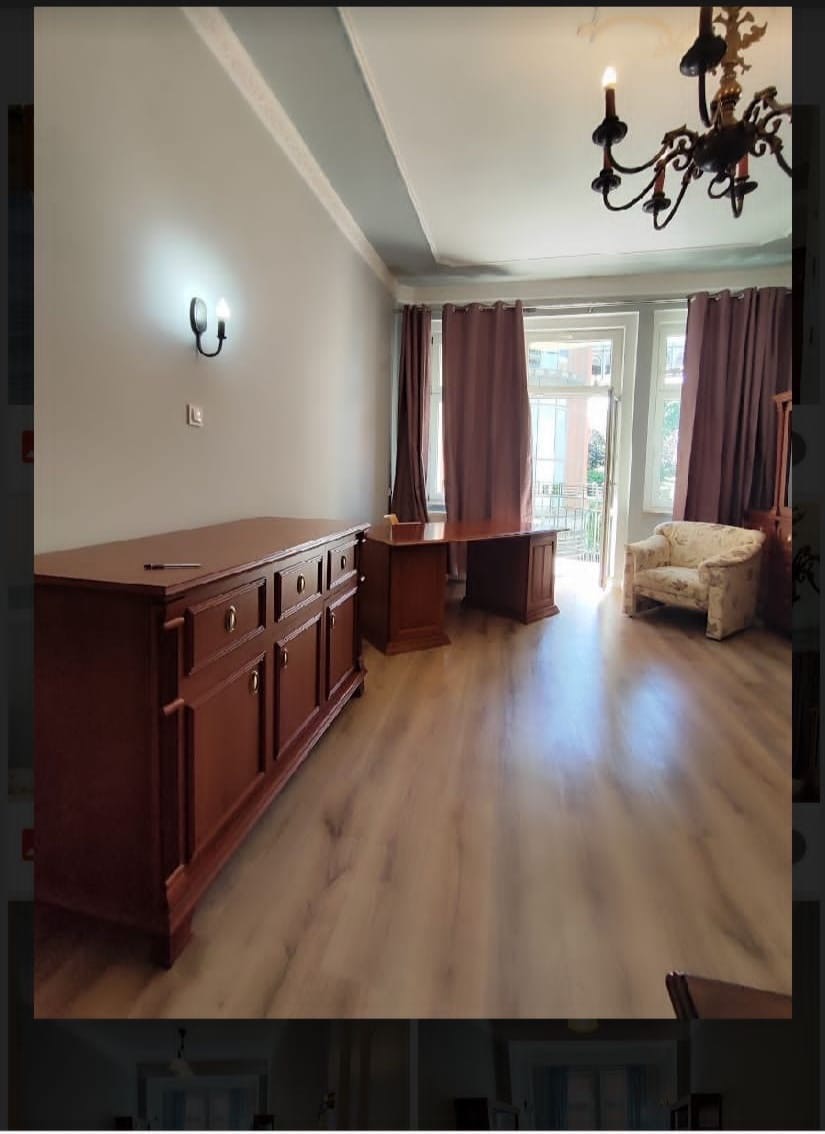 Jasne przestronne wnętrze z drewnianą komodą, biurkiem i ozdobnym fotelem, oświetlone żyrandolem i kinkietem, z widokiem na balkon.