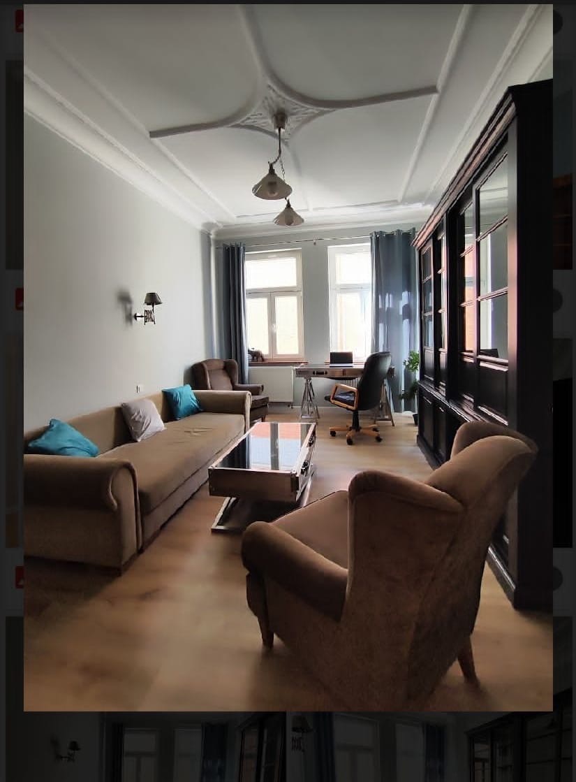 Jasne, przestronne wnętrze salonu z elementami biura: sofa, fotel, stolik kawowy, biurko z krzesłem, regał z przeszkleniami, sztukateria na suficie, drewniana podłoga.