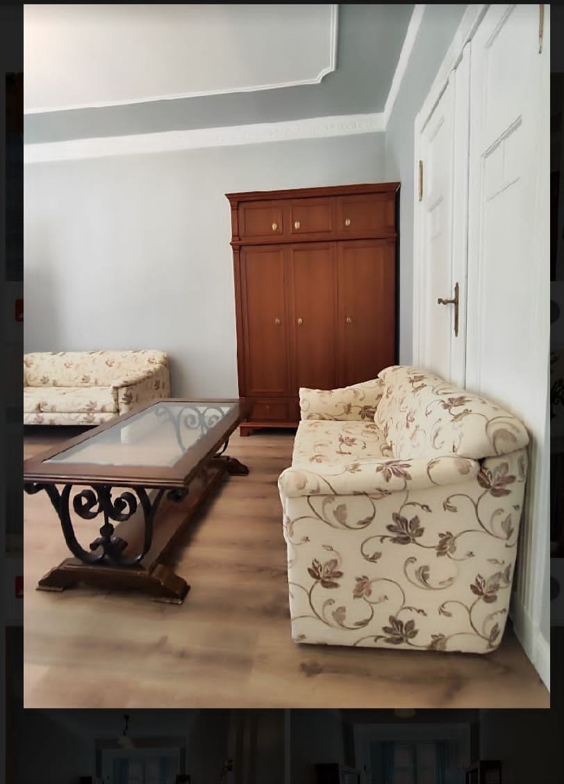 Umeblowany salon z drewnianą szafą, sofą w kwiaty i stolikiem kawowym z metalowymi nogami o dekoracyjnym wzorze.