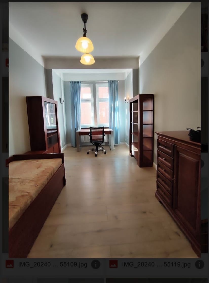 Pokój z drewnianą podłogą, biurkiem przy oknie z niebieskimi zasłonami, regałami i komodą w stylu retro.