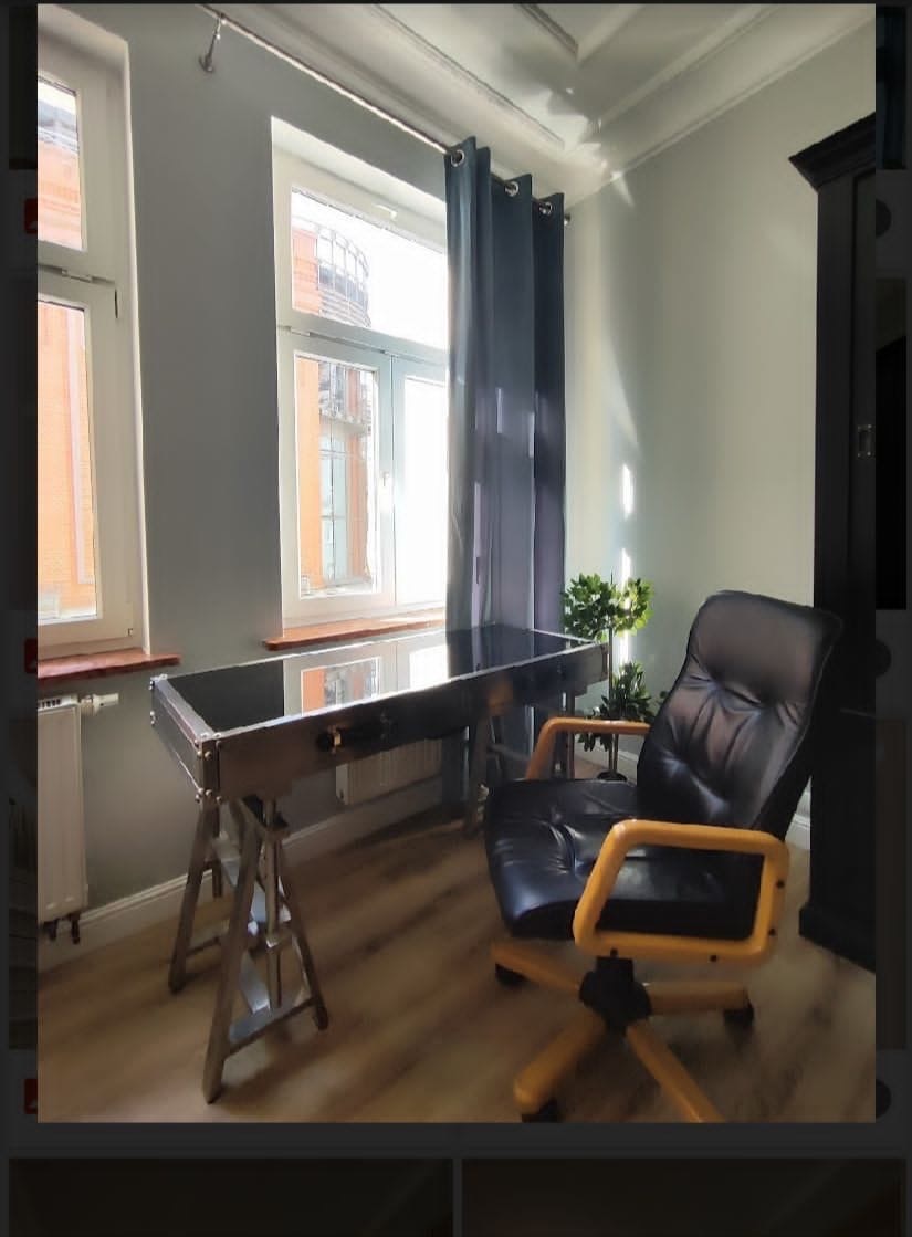 Pokój z biurkiem o metalowej konstrukcji z blatem z ciemnego szkła i czarnym skórzanym krzesłem biurowym na drewnianej podłodze, widok na okno z szarymi zasłonami.