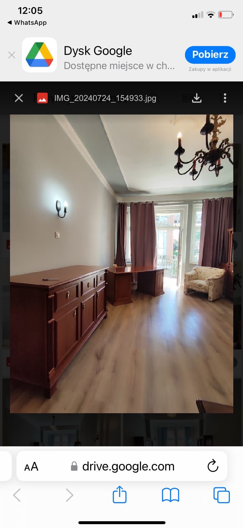 Jasne wnętrze pokoju z drewnianą podłogą, brązowymi meblami w stylu retro, w tym komodą i biurkiem, oraz kremowym fotelem w kwiaty, oświetlone przez czarny żyrandol i kinkiet na ścianie