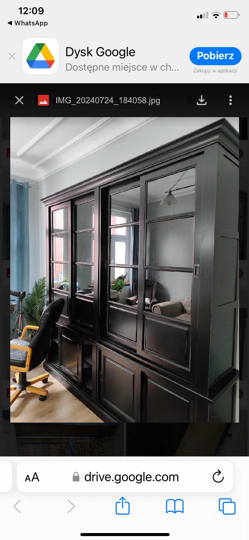 Czarny, drewniany regał z przesuwanymi, przeszklonymi drzwiami w jasnym pomieszczeniu. Widoczny fotel biurowy i elementy dekoracyjne wewnątrz.