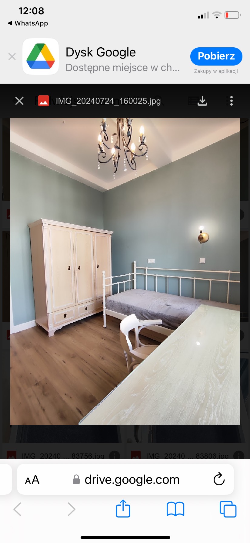 Jasne wnętrze sypialni z drewnianą podłogą, pomalowanymi na zielono ścianami, kremową szafą, metalowym łóżkiem z szarą narzutą, białym krzesłem i fragmentem biurka, oświetlone żyrandolem...