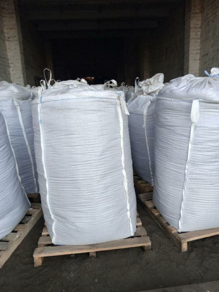 Magazynowane, białe worki typu big bag z zawartością na drewnianych paletach w słabo oświetlonym wnętrzu budynku.