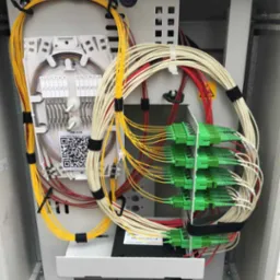 Szafka telekomunikacyjna z uporządkowanymi kablami światłowodowymi w kolorach żółtym, białym i czerwonym, zakończonymi zielonymi złączkami, widoczny panel krosowniczy z numeracją.