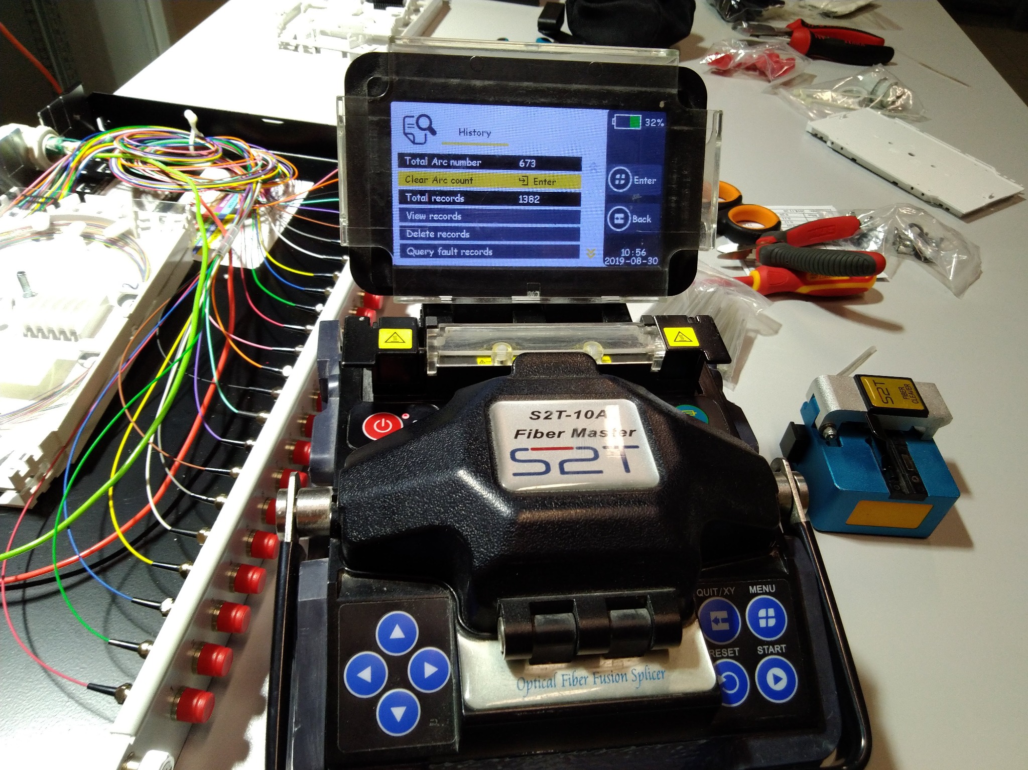 Zgrzewarka światłowodowa S2T-10A Fiber Master z widocznym ekranem historii i statystyk zgrzewów, obok splitter światłowodowy z kolorowymi kablami oraz narzędzia na białym stole.