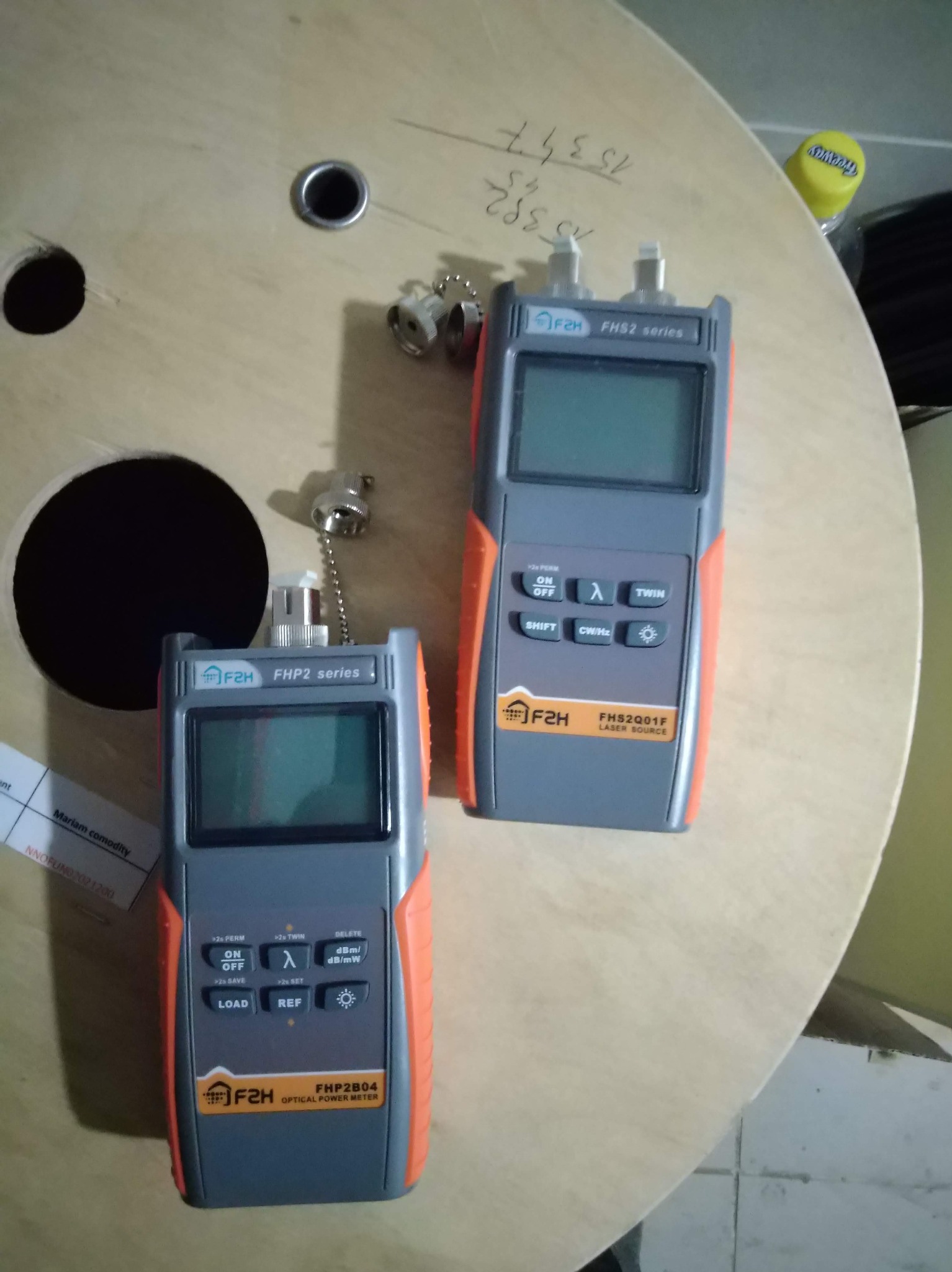 Dwa mierniki optyczne F2H (model FHP2B04 i FHS2Q01F) leżące na drewnianej szpuli kablowej, jeden z podłączonym złączem światłowodowym.