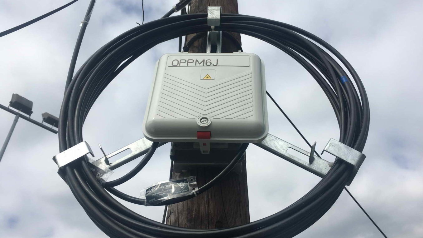 Skrzynka przyłączeniowa OPM6J zamontowana na słupie drewnianym, otoczona pętlą kabli. Widoczne metalowe uchwyty mocujące i fragment nieba w tle.